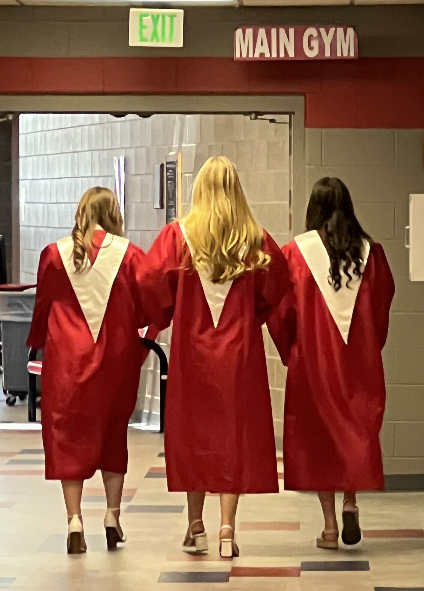 three female students with long hair, walking into the main gym at Platteview high school, wearing their gowns for graduation